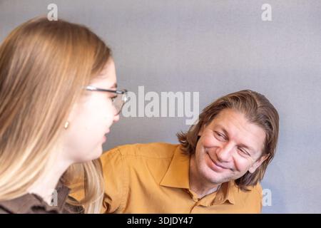 Middle-aged man interacts with a young woman in glasses, both positioned closely and facing each other in a neutral gray setting Stock Photo