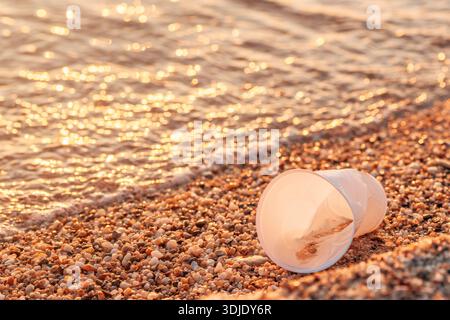 Seaside pollution. Plastic cup on a pebble beach at sunset, with golden reflections on the water and shoreline. Selective focus. Stock Photo