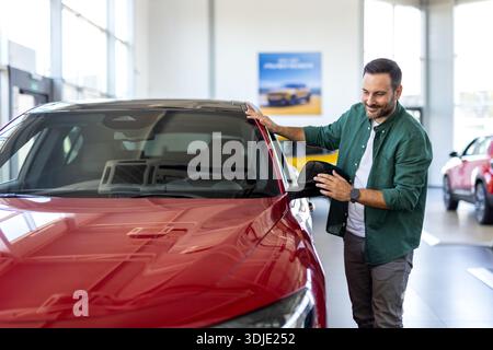 Handsome man buying new car, standing at car salon and checking automobile surface. Smiling guy looking at beautiful red auto in showroom Stock Photo