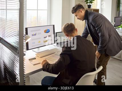 Two businessmen analyzing finance charts on computer in modern office workspace Stock Photo