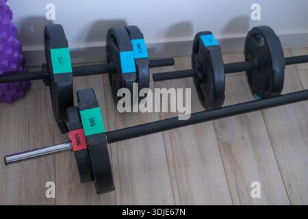 Diagonal close-up of black adjustable dumbbell and barbell with coloured weight markers on laminate floor; white wall and skirting board Stock Photo