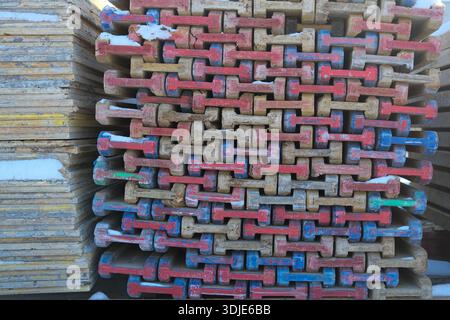 Tight front view of stacked wood formwork with chipped red and blue paint on end blocks; adjacent plywood sheets; snow patches; crisp daylight shadows Stock Photo