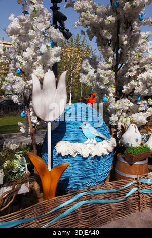 Colorful Easter decoration with a large blue egg, wooden tulip, and bird, set among cherry blossom trees in a vibrant outdoor environment. Stock Photo