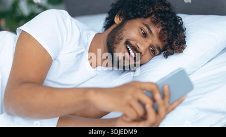 Man Lying on Bed Looking at Smartphone and Smiling While Enjoying Content Stock Photo
