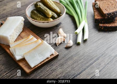Pork fatback on a wooden cutting board with slices of rye bread, garlic, pickled cucumbers in a plate and fresh green onions, closeup Stock Photo