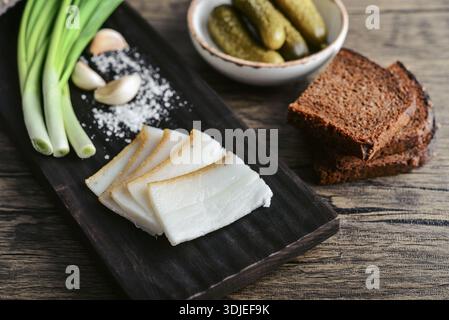 Pork fatback on a wooden cutting board with slices of rye bread, garlic, pickled cucumbers in a plate and fresh green onions, closeup Stock Photo