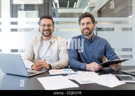 Two cheerful male professionals sitting together at a desk in a contemporary office, reviewing market reports and documents while working on a laptop, embodying teamwork and successful partnership Stock Photo