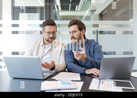 Two business professionals intensely collaborating at a conference table, reviewing laptops and documents while planning strategy and brainstorming solutions in a modern office setting Stock Photo