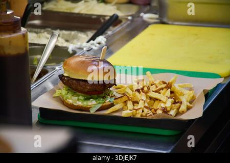 Close view of handcrafted hamburger and crispy fries prepared by cook wearing gloves in modern food service environment, quality ingredients, street f Stock Photo