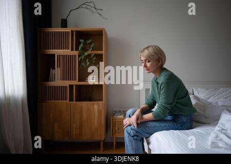 Pondering lonely woman sits on bed with upset expression, coping with thoughts about life crisis. Stock Photo
