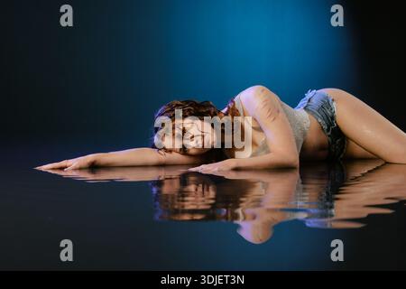 Female contemporary dancer lies on wet reflective floor in studio light with copy space. Ideal for banner or website about dance classes, performance, Stock Photo