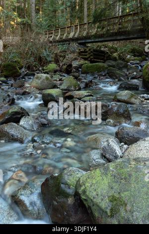 A stream of water flows between large rocks. The water is clear and the rocks are grey. The scene is peaceful and serene. There is a wooden footbridge Stock Photo