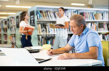 Focused casual mature male writing in notepad and reading books in the library Stock Photo