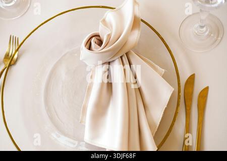 Beautifully arranged table setting showcases a clear glass plate with golden rim, cream napkin, golden cutlery, and crystal glasses, ideal for elegant Stock Photo