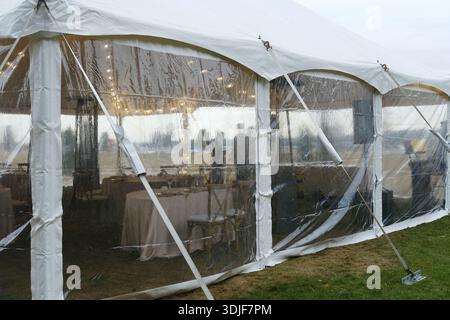 Clear tent with white fabric and transparent sides, tables with white tablecloths inside, outdoor grass area with cloudy sky and distant figures visib Stock Photo