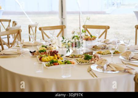 Round table with white tablecloth features plates of food, glasses, cutlery, and a floral centerpiece with various dishes arranged on it in a tent set Stock Photo
