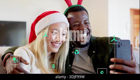 Smiling adult couple taking selfie in living room, with smartphone, Santa hat, antlers, green icons Stock Photo