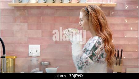 Holding green mug near mouth, redhead in fluffy-knit sweater at kitchen counter, copy space Stock Photo