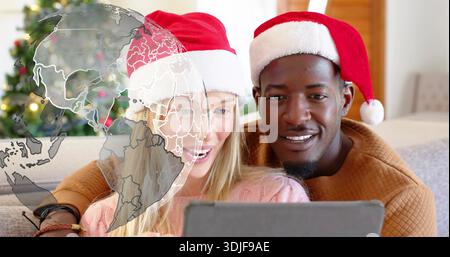 Holding tablet couple sitting on sofa wearing Santa hats pink sweater brown knit globe overlay tree Stock Photo