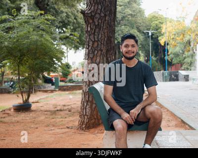 Photograph of a young man sitting in an urban park setting, smiling and looking at camera, with trees and outdoor areas in the background. Useful for Stock Photo