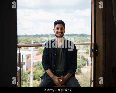 Image of a young man sitting on a balcony or terrace, smiling at the camera, with a view of the city and sky in the background. Perfect for communicat Stock Photo