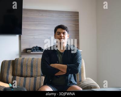 Indoor photo of a young man sitting on a sofa with his arms crossed, looking at camera in a living room. Useful for content from everyday life, home, Stock Photo