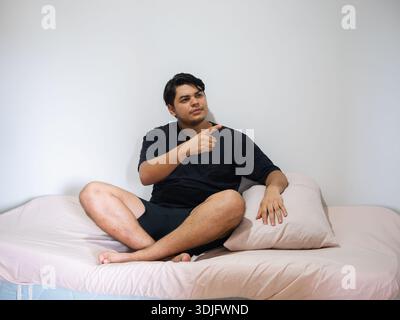 Indoor image of a young man sitting on a bed, pointing to the side, in a bedroom with a clean background. Useful for advertisements with space for cop Stock Photo