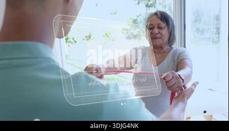 Performing senior woman holding red resistance band by window, assistant in teal shirt, hologram Stock Photo