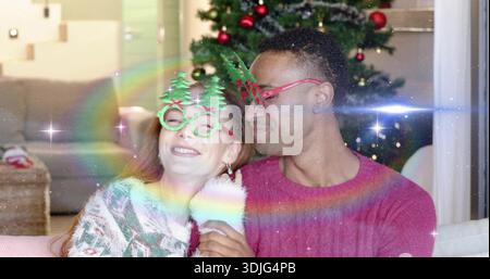 Smiling couple on home couch, wearing sweaters and novelty glasses, woman holding fuzzy mug by tree Stock Photo