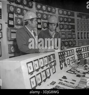 Sloviansk, Ukraine, USSR - 1980s: Two engineers in safety helmets monitor the production process at the main control panel of a large industrial enterprise. One specialist checks the readings of analog gauges on the wall while his colleague records data in a logbook. This authentic black and white archival photograph captures the nerve center of a Soviet factory, possibly a power station or a heavy industry plant. The image documents the technical expertise and management of complex systems in the peaceful Donbas region long before the war. Stock Photo