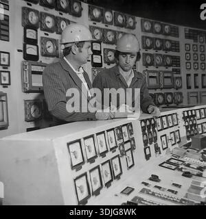 Sloviansk, Ukraine, USSR - 1980s: Two engineers in safety helmets monitor the production process at the main control panel of a large industrial enterprise. One specialist checks the readings of analog gauges on the wall while his colleague records data in a logbook. This authentic black and white archival photograph captures the nerve center of a Soviet factory, possibly a power station or a heavy industry plant. The image documents the technical expertise and management of complex systems in the peaceful Donbas region long before the war. Stock Photo