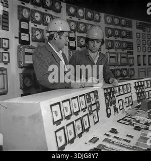Sloviansk, Ukraine, USSR - 1980s: Two engineers in safety helmets monitor the production process at the main control panel of a large industrial enterprise. One specialist checks the readings of analog gauges on the wall while his colleague records data in a logbook. This authentic black and white archival photograph captures the nerve center of a Soviet factory, possibly a power station or a heavy industry plant. The image documents the technical expertise and management of complex systems in the peaceful Donbas region long before the war. Stock Photo