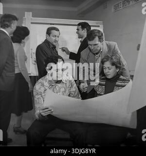 Sloviansk, Ukraine, USSR - 1980s: A team of engineers and designers working in the design bureau of a Soviet industrial plant. A young specialist in a patterned shirt holds a large paper blueprint while a senior colleague points out technical details to a female employee. In the background, other employees discuss a project at a drawing board stand. This authentic black and white archival photograph captures the intellectual labor and the engineering planning process in the peaceful Donbas region long before the war. Stock Photo
