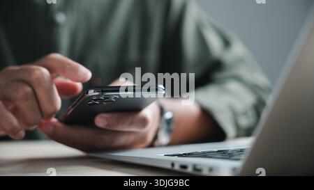 Close-up of a person's hand holding a smartphone with a laptop in the background. The image focuses on the smartphone's camera lens. Stock Photo