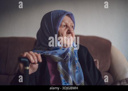 Portrait of an old thoughtful Muslim woman with her walking stick at home wearing traditional hijab, looking away. Stock Photo
