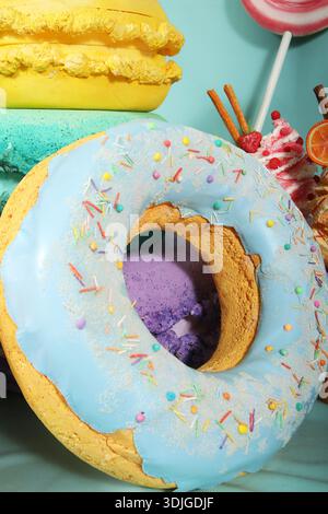 Large artificial donut with blue icing and colorful sprinkles, surrounded by various sweets on a turquoise background, creating a fun and whimsical de Stock Photo