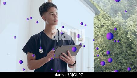 Standing male medic checking tablet on home porch, in navy scrubs, stethoscope, purple spheres Stock Photo