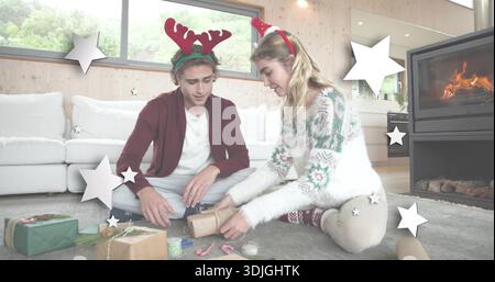 Wrapping friends in reindeer headbands and holiday sweaters on rug at home, with gift wrap supplies Stock Photo
