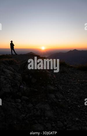 A hiker silhouetted against the sunrise on the summit of the Geigelstein in the Chiemgau Alps, Bavaria, Germany Stock Photo