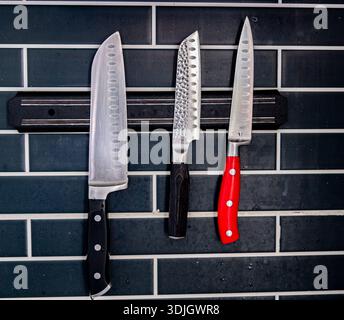 Three kitchen knives hanging on a tiled wall. Stainless steel blades with different handles, modern kitchen interior detail. Stock Photo