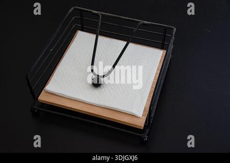 Stack of square paper napkins held in a metal napkin holder on a dark surface. Minimal still life composition with neutral background. Stock Photo