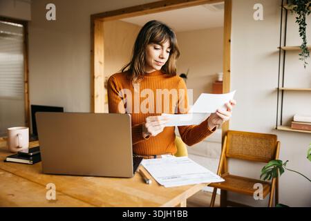 Woman managing finances, checking documents, and using laptop at wooden desk Stock Photo