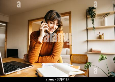 Woman smiling and talking on smartphone, working from home on laptop Stock Photo