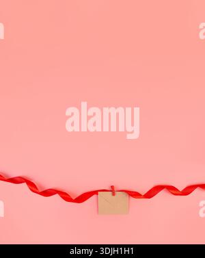 craft envelope attached to a swirling red ribbon isolated on a pastel pink background with copy space. Object for design to women's day, mother's day, Stock Photo