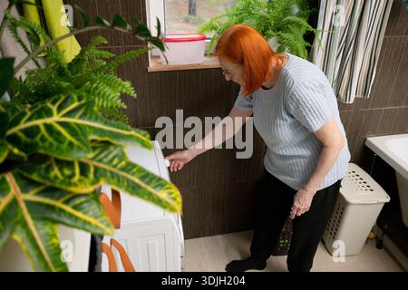 A detailed overhead perspective showing a senior woman's hands adjusting the settings on a shiny silver washing machine before starting it. Stock Photo