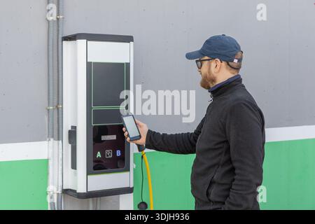 A young man charges an electric car at an outdoor charging station, using a smartphone app to monitor and control the process, with a cable connected Stock Photo