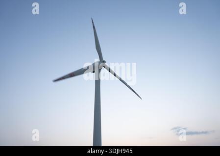 A single large wind turbine stands tall against a clear sky with its blades slowly turning, generating clean energy. Stock Photo