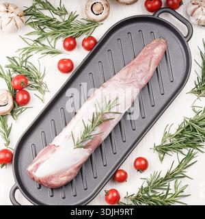 A raw pork tenderloin rests on a black cast iron grill pan, garnished with fresh rosemary sprigs and scattered cherry tomatoes. This image captures th Stock Photo