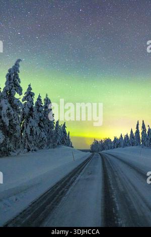 View of a snowy road cutting through a winter wonderland beneath the ethereal glow of the Northern Lights, Pudasjärvi, North Ostrobothnia, Finland. Stock Photo