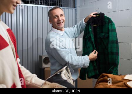 Middle aged Caucasian man smiling while holding green plaid jacket on hanger, standing in storage room with young adult man handing over pair of ice skates, engaging in sorting or donation activity Stock Photo